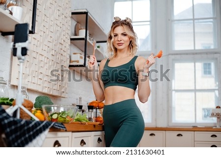 Side view of female fitness blogger with slender body recording video on smartphone while cooking fresh salad on kitchen. Concept of healthy diet and sport nutrition.