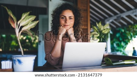 Portrait of millennial female IT professional with modern laptop device looking at camera during time for creating graphic design, skilled woman with digital netbook posing at terrace table