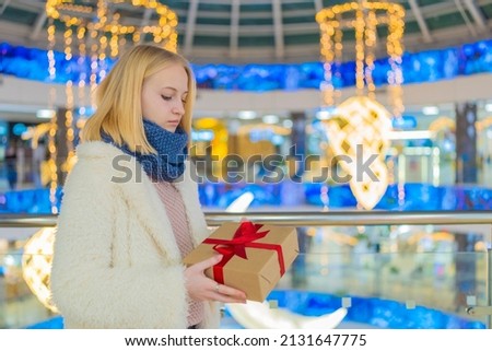 teenage girl in a white jacket in a mall, holding a box with a gift on the background of a shop window with discounts during a sale.