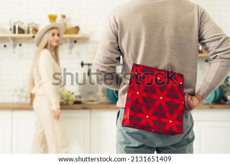 European man standing and holding red gift bag behind his back for his long-haired blonde partner who is standing in the kitchen. High quality photo