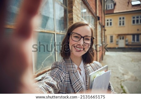Young smiling business woman with paperwork, documents and a laptop in hand taking a selfie outside in the nice weather with old buildings in the background. Concept: businesswoman or female coworker 