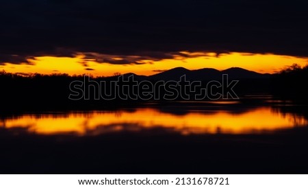 Last light in sky above mountain silhouette framed with dark clouds at twilight - landscape with symmetric reflection on water surface