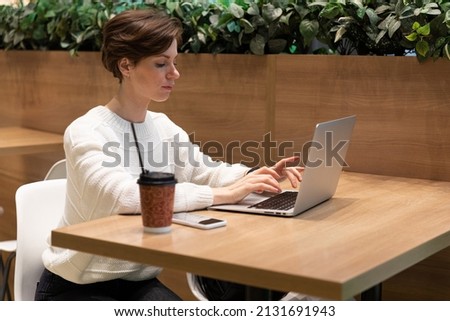 A photo of a young woman who sits at the food court of a shopping center at a table and works at a laptop. Freelance concept