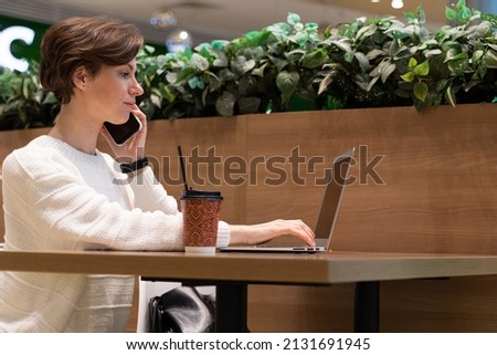 A photo of a young woman who sits at the food court of a shopping center at a table and works at a computer laptop, talks on a mobile phone. Freelance concept