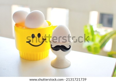 Happy Easter Day celebration concept, cute yellow happy face mug put many white eggs and white ceramic cup of one egg on white table blurred corridor background, symbolic holiday season concept