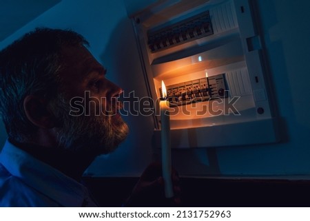 Energy crisis. Man in complete darkness holding a candle to investigate a home fuse box during a power outage. Blackout concept. Royalty-Free Stock Photo #2131752963