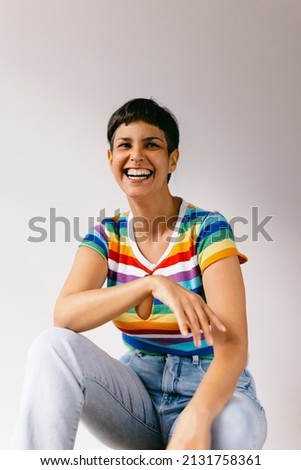 Lesbian woman laughing cheerfully during pride month. Happy young lesbian woman celebrating gay pride against a studio background. Young woman wearing a tank top with LGBTQ+ rainbow colours. Royalty-Free Stock Photo #2131758361