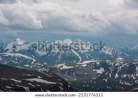 Awesome aerial view to high snow mountains under cloudy sky. Gloomy mountain landscape at very high altitude with cloudiness. Dark atmospheric mountain scenery with snowy mountain range in overcast.