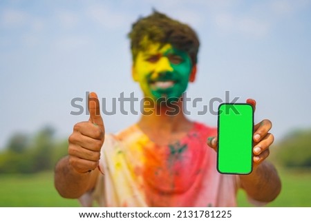 Young Indian boy covered in holi colors holding a mobile phone with green screen showing thumbs up. Holi festival and technology concept.