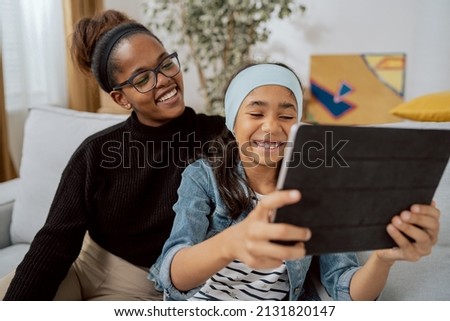 A beautiful girl and her elegant mom with glasses are sitting in an apartment on the couch, languishing, the daughter is holding a tablet in hands, taking pictures, selfie with mother on social media