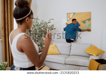 Couple in love decorating their first apartment, unpacking boxes after moving in, the man hanging a painting on the wall, the woman showing how much to lift up to make it even