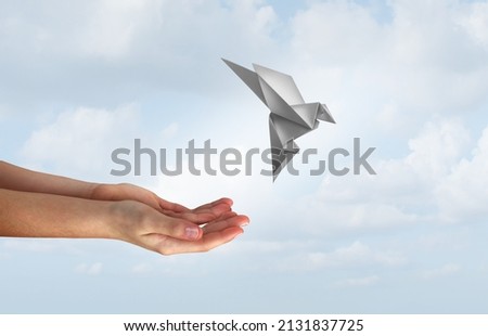 Concept of hope and freedom as human hands release a magical flying origami bird made of paper as a liberty and democracy symbol for peace and love of humanity to stop war and spirituality icon. Royalty-Free Stock Photo #2131837725