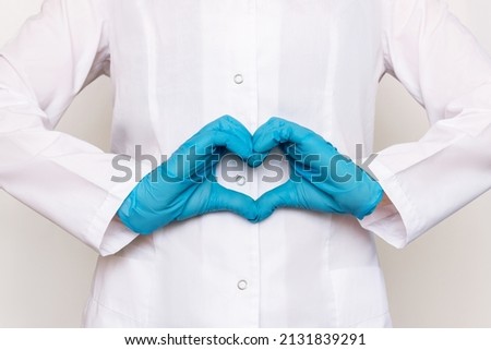 Cropped shot of a young female doctor in a white coat forming a heart shape with her hands in blue gloves isolated on a gray background