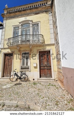 Yellow tile facade of old Neoclassical townhouse with black bicycle stopped at the door on the cobblestone sidewalk and chipped wooden door under metal railing balcony. Tavira-Algarve region-Portugal. Royalty-Free Stock Photo #2131862127