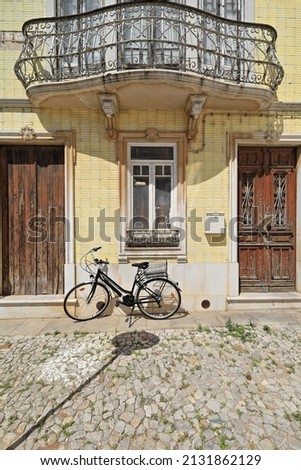 Yellow tile facade of old Neoclassical townhouse with black bicycle stopped at the door on the cobblestone sidewalk and chipped wooden door under metal railing balcony. Tavira-Algarve region-Portugal. Royalty-Free Stock Photo #2131862129