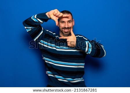 Cheerful funny man in casual clothes posing isolated on blue wall background, studio portrait. People candid emotions lifestyle concept. Mock up copy space. Make hands photo frame gesture.