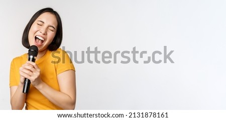 Happy asian girl dancing and singing karaoke, holding microphone in hand, having fun, standing over white background