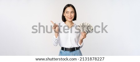 Smiling young modern asian woman, pointing at banner advertisement, holding cash money dollars, standing over white background