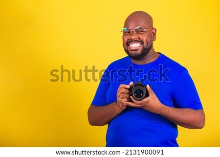 handsome afro brazilian man wearing glasses, blue shirt over yellow background. holding photo camera, photographer, smiling, creative. DSLR