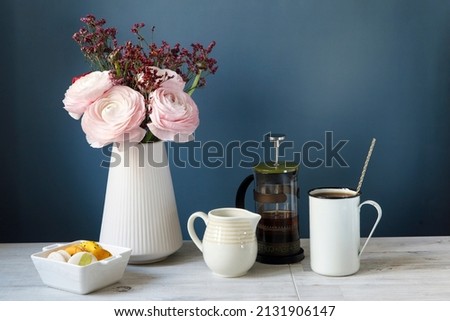 Coffee cup and bouquet of pink Persian buttercups on wooden table. mock up background for design. Yellow and white macaroons in a white porcelain bowl. Dark blue background. Copy space
