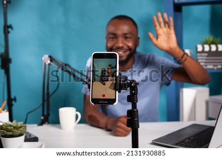 Selective focus on mobile phone recording social media influencer waving hello sitting at desk with professional microphone. Closeup of live vlog setup with smarthone on stand filming vlogger.
