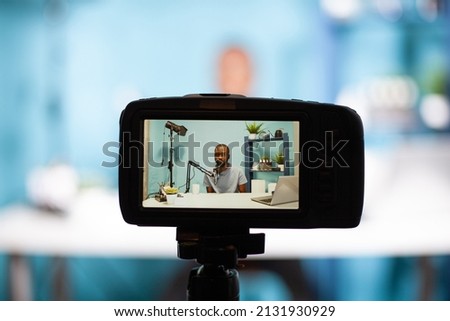 Closeup of professional digital camera on tripod recording vlogger talking at professional microphone in vlogging studio. Selective focus on back screen of dslr filming influencer sitting at desk.