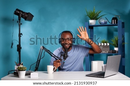 Smiling influencer wearing wireless headphones waving hello in recording studio with professional microphone and video light. Happy vlogger doing hand gesture in front of audio podcast setup.