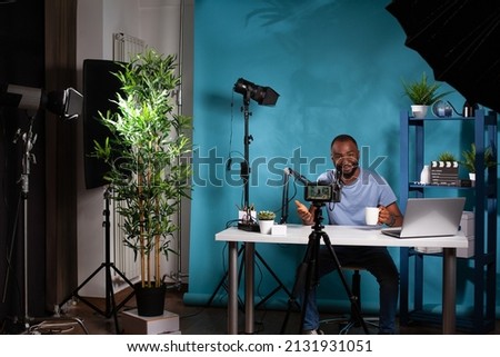 Content creator talking to followers in in front of filming video camera on tripod sitting at desk holding cup. Wide view of vlogging studio setup with smiling vlogger interacting with audience.