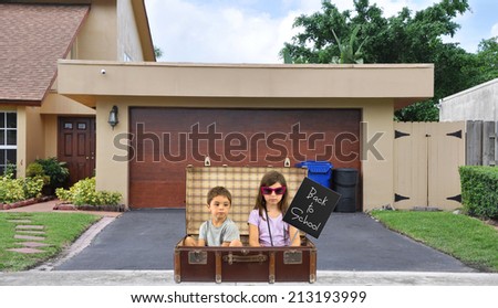 Back to School Children sitting in vintage suitcase on blacktop driveway of suburban home residential neighborhood USA blue sky clouds
