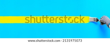 Hand holding paintbrush painting stripe yellow color on blue wall. Colorful art brush strokes painted for home improvement decoration. Horizontal banner backdrop with copy space of Ukraine concept. Royalty-Free Stock Photo #2131975073