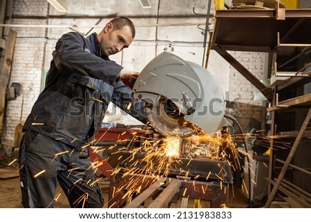 A young man in shabby work clothes, gloves, cuts metal without wearing goggles on a miter saw in violation of safety regulations in the workshop of an industrial enterprise. Sparks fly. Royalty-Free Stock Photo #2131983833