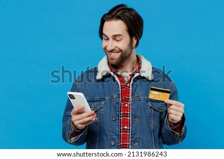 Young brunet bearded man 20s in denim jacket use mobile cell phone hold credit bank card do online shopping isolated on plain pastel light blue background studio portrait. Winter cold season concept