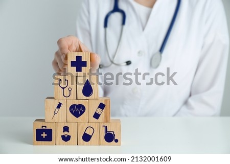 Doctor building pyramid of wooden cubes with different icons at white table, closeup. Insurance concept