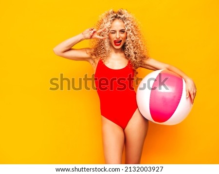 Young beautiful smiling woman posing near yellow wall in studio.Sexy model in red swimwear bathing suit.Positive female with curls hairstyle. Holding penny inflatable ball.Happy and cheerful Royalty-Free Stock Photo #2132003927