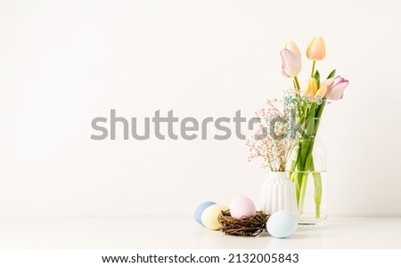 Home interior with easter decor. Fresh spring tulips in vase, hypsophila and pastel colored eggs on white wall background, copy space
