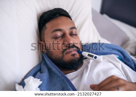 A sick man lies with a thermometer reading 38.3 degrees Celcius for a fever. High quality photo Royalty-Free Stock Photo #2132027427
