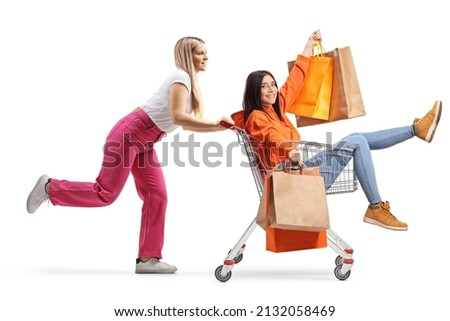 Young female pushing her friend inside a shopping cart with shopping bags isolated on white background