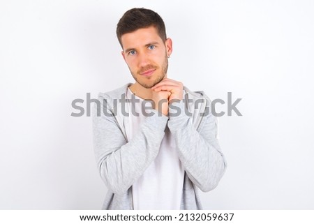 Charming serious young caucasian man wearing casual clothes over white background keeps hands near face smiles tenderly at camera
