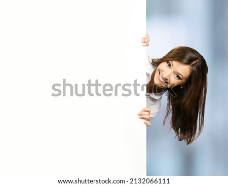 Happy smiling brunette young woman, standing behind, peeping from blank banner or mock up signboard, copy space area for text, indoors. Mockup ad advertisement billboard bill board big big board.