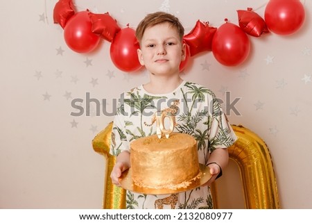 A ten-year-old boy in red shorts holds a large gold cake. birthday.red balloons
