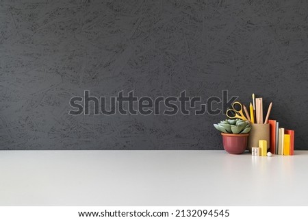 Home office desk with school or office supplies, books and plant on a black wall background.