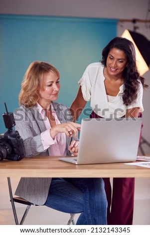 Mature Female Fashion Photographer With Client Looking At Images From Shoot On Laptop In Studio