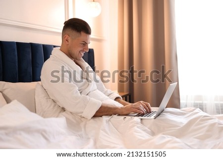 Handsome man wearing bathrobe with laptop on bed in hotel room
