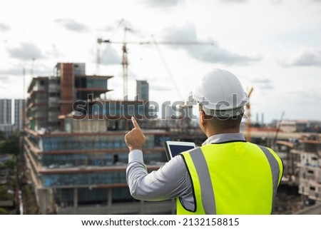 Asian engineer handsome man or architect use tablet with white safety helmet in city construction site . Standing on rooftop building construction at capital. Royalty-Free Stock Photo #2132158815
