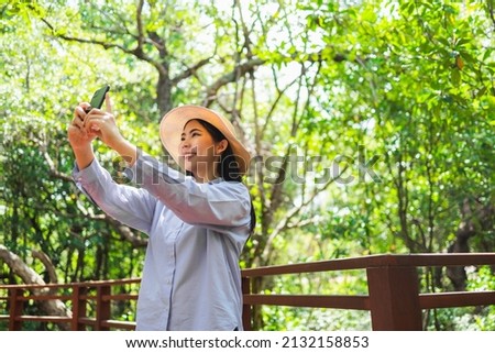 Asian woman travel using cell phone taking video and photo in nature with sunlight. Traveller blogger in mangrove forest. Rest on vacation holiday weekend. Beautiful female with hat and blue shirt