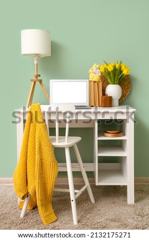Workplace with laptop, tulips and Easter rabbit with greeting card near color wall in room interior