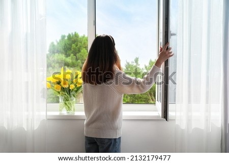 Pretty young Asian woman opening window at home on sunny day, back view Royalty-Free Stock Photo #2132179477