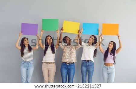 Group of happy smiling young multiracial women looking up at colorful purple, green, yellow, blue and orange mockup paper sign banners they're holding standing together against white grey studio wall