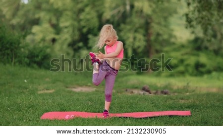 Ukrainian beautiful 7 year old girl with white hair like an angel is engaged in fitness in green field near the river.A sporty girl does gymnastic exercises on a mat.A healthy lifestyle.