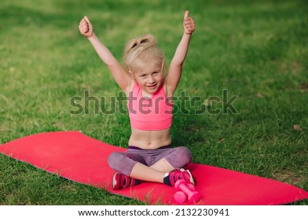 Ukrainian beautiful 7 year old girl with white hair like an angel is engaged in fitness in green field near the river.A sporty girl does gymnastic exercises on a mat.A healthy lifestyle.